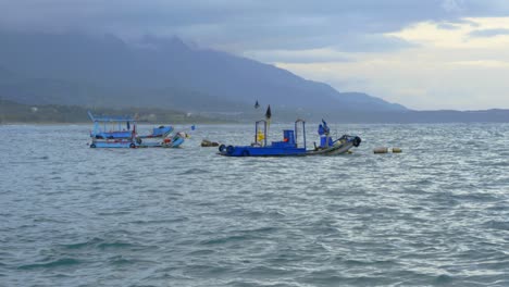 Vor-Anker-Liegende-Asiatische-Fischerboote-Schaukeln-Im-Sanften-Wellengang-Des-Ozeans-Mit-Bergen-Und-Bewölktem-Himmel-Im-Hintergrund,-Gefilmt-Als-Stationäre-Halbtotalaufnahme