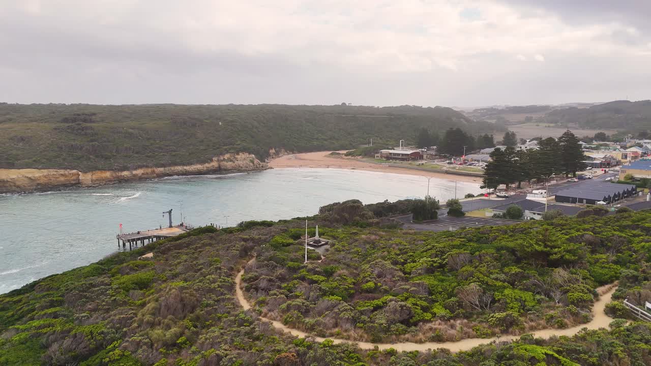 Drone footage captures Port Campbell's rugged coastline, showcasing cliffs, ocean waves, and a small town under overcast skies