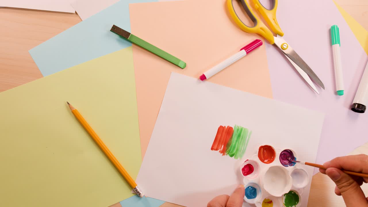 Hand uses brush to paint colorful stripes on paper, surrounded by stationery, under soft lighting
