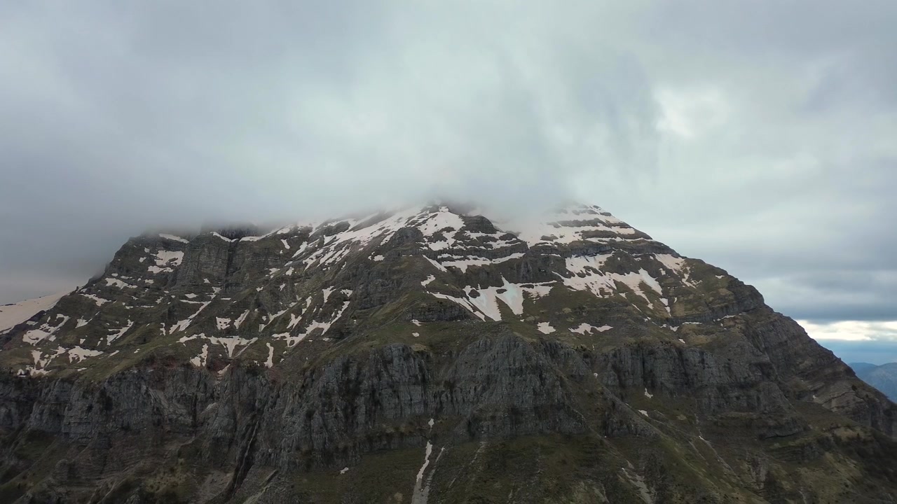 Cinematisk 4K droneoptagelse med langsom udzoomning, der afslører de barske, sneklædte klipper på Stroggoula-toppen, indhyllet i stemningsfulde stormskyer. Episk atmosfærisk alpint vildnis i Epirus.