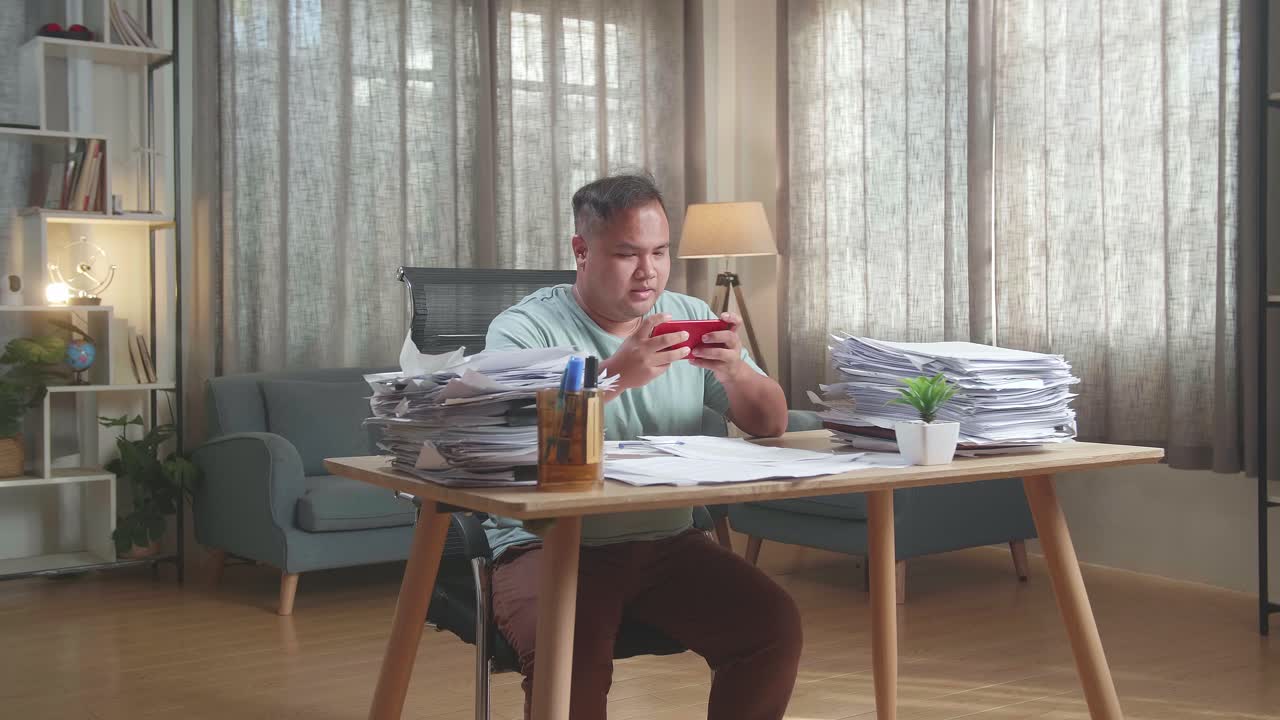 Fat Asian Man Playing Game On Smartphone After Working With Documents At The Office