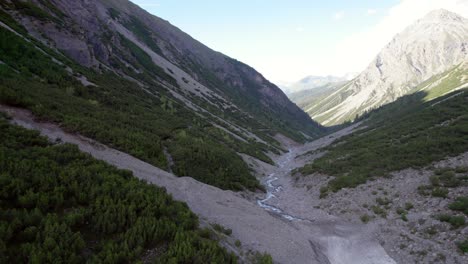 Imágenes-Aéreas-De-Drones-Volando-A-Lo-Largo-De-Las-Laderas-De-Las-Montañas-A-Través-De-Un-Espectacular-Valle-Glacial-Rodeado-De-Pinos-Con-Parches-De-Nieve-Y-Un-Río-Alpino-En-Suiza