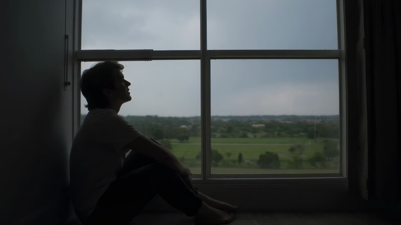Sitting man at home in tee looking at fields, backlight casting silhouette at window, copy space