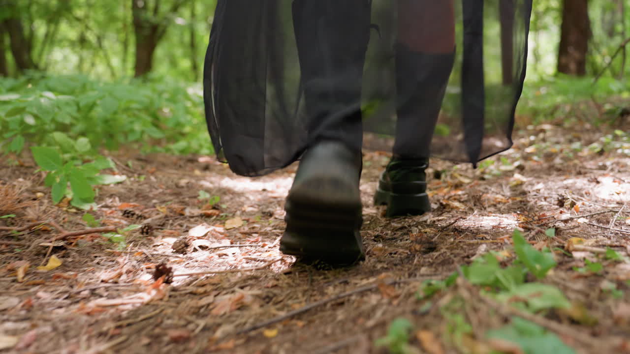 Rear lower angle view of warrior walking through forest holding wooden staff, long robe swaying and boots stepping on earthy ground surrounded by green leaves