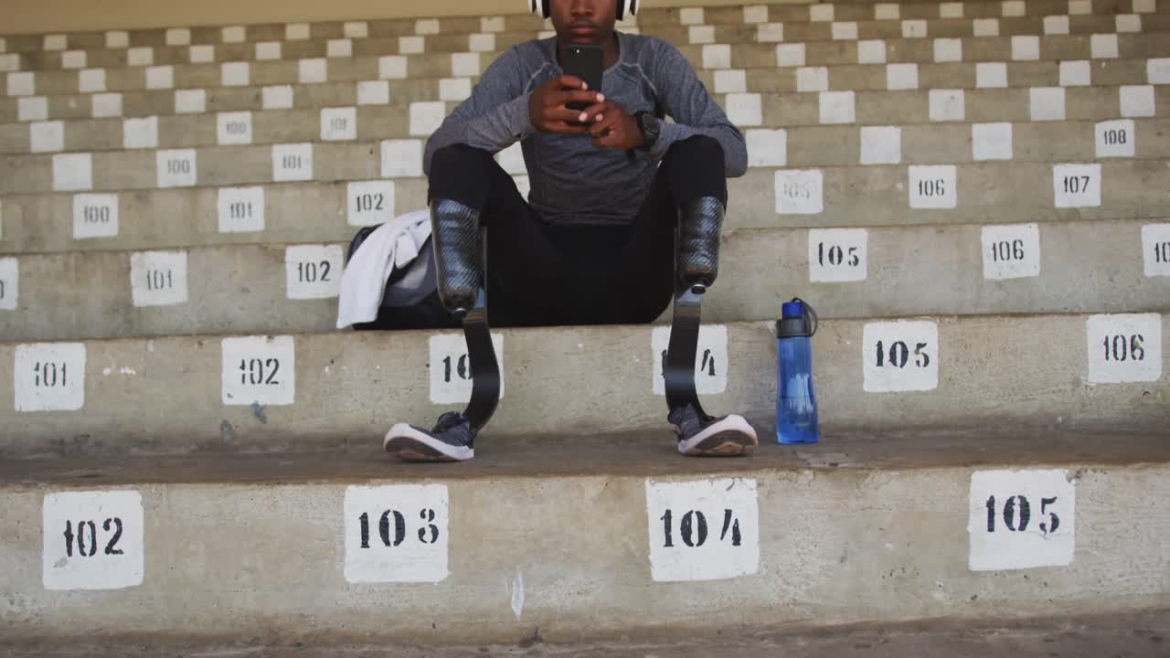 Disabled mixed race man with prosthetic legs sitting on a stadium and using a smartphone