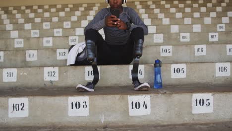 disabled mixed race man with prosthetic legs sitting on a stadium and using a smartphone