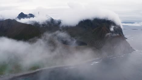 Aerial-view,-pushing-through-the-clouds-on-a-moody-day,-revealing-a-gorgeous-lake-resting-in-the-mountains-above-the-sea,-near-Unstad,-Lofoten-Islands,-Norway