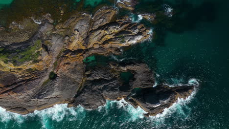 Aerial:-waves-crashing-against-rocky-islet-in-clear-turquoise-sea-water