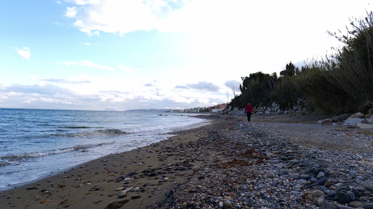 vista de una persona caminando a lo largo de la playa de grava de piedra de estepona con pequeñas olas rompiendo