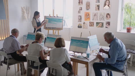 High-angle-view-of-cheerful-senior-friends-painting-on-canvas.-Senior-woman-smiling-while-drawing-with-the-group.-Seniors-Attending-Painting-Class-Together.-Senior-men-having-fun-painting-in-art-class