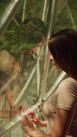 young lady looks out window examining area with dense forest. sun rays fall on female with reflection in panoramic window of modern apartment for tourists upper view