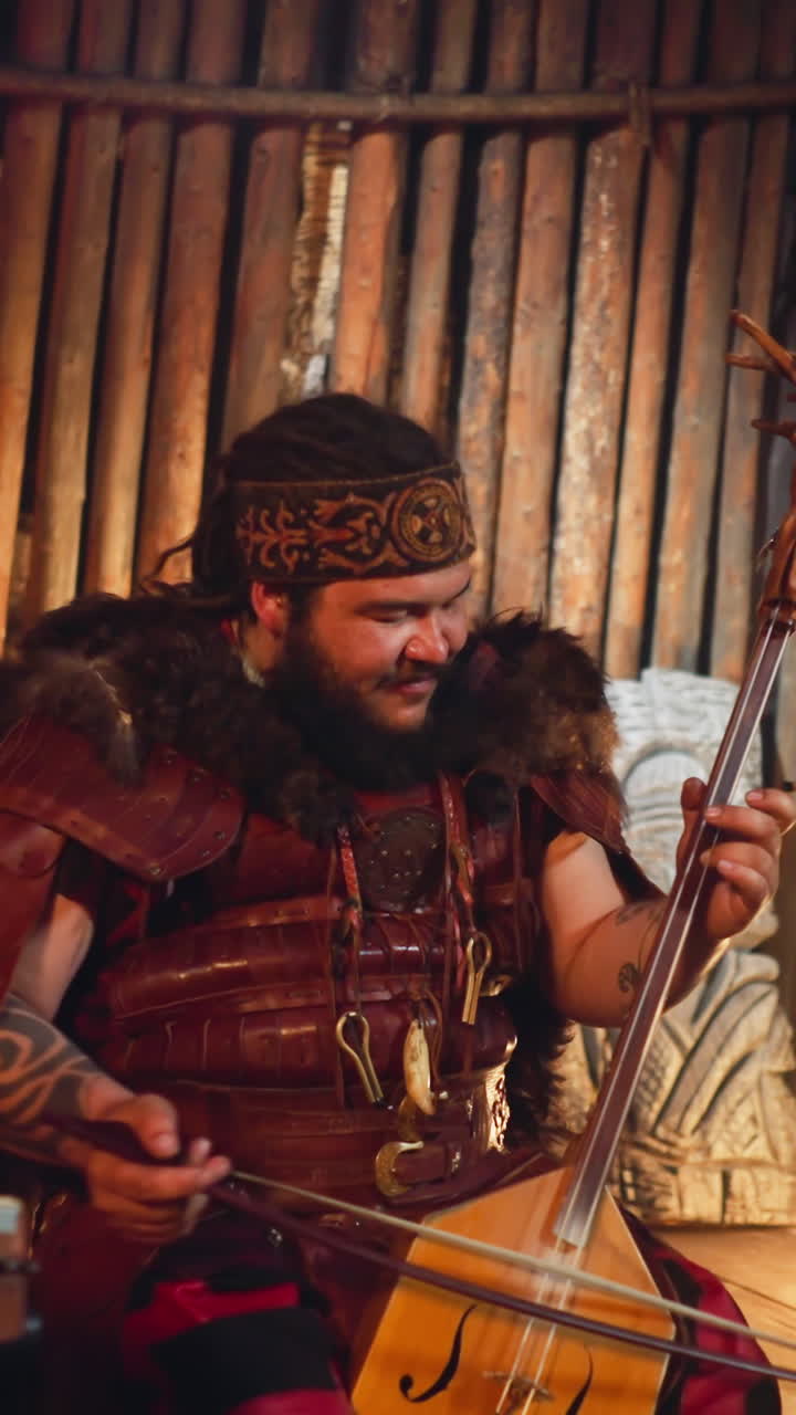 Obese man in Altai ethnic clothes plays bow instrument decorated with tree roots and drummer on stage. Encouraged performing of old Siberian culture