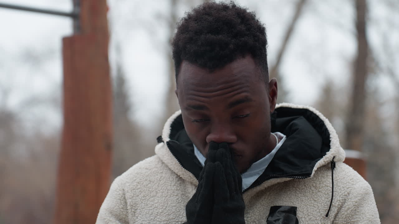 Man met zwarte handschoenen en een witte winterjas buiten in koud weer, handen samengevouwen bij het gezicht, peinzende uitdrukking met wazige bomen en structuren op de achtergrond.