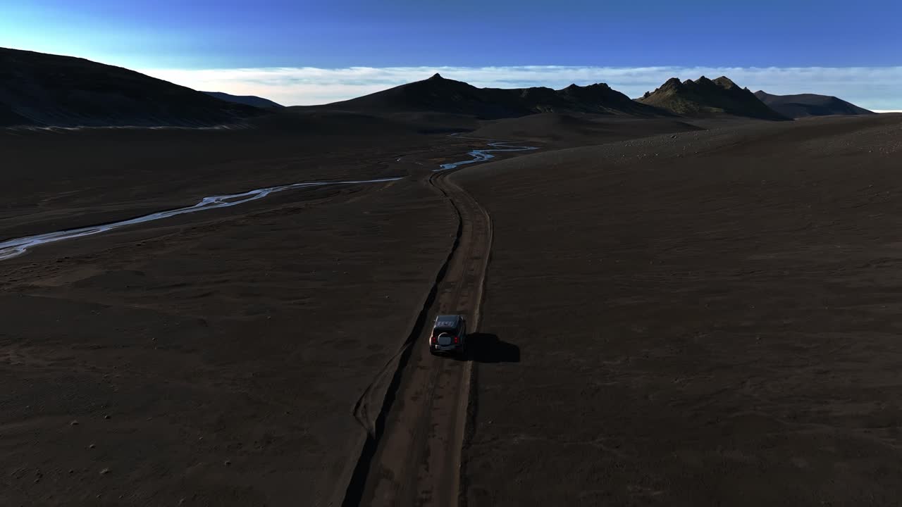 Ripresa aerea con drone di un SUV 4x4 che percorre una pista stretta attraverso gli altopiani islandesi di sabbia nera, con canali tortuosi di acqua di disgelo e distanti picchi vulcanici sotto un cielo blu.