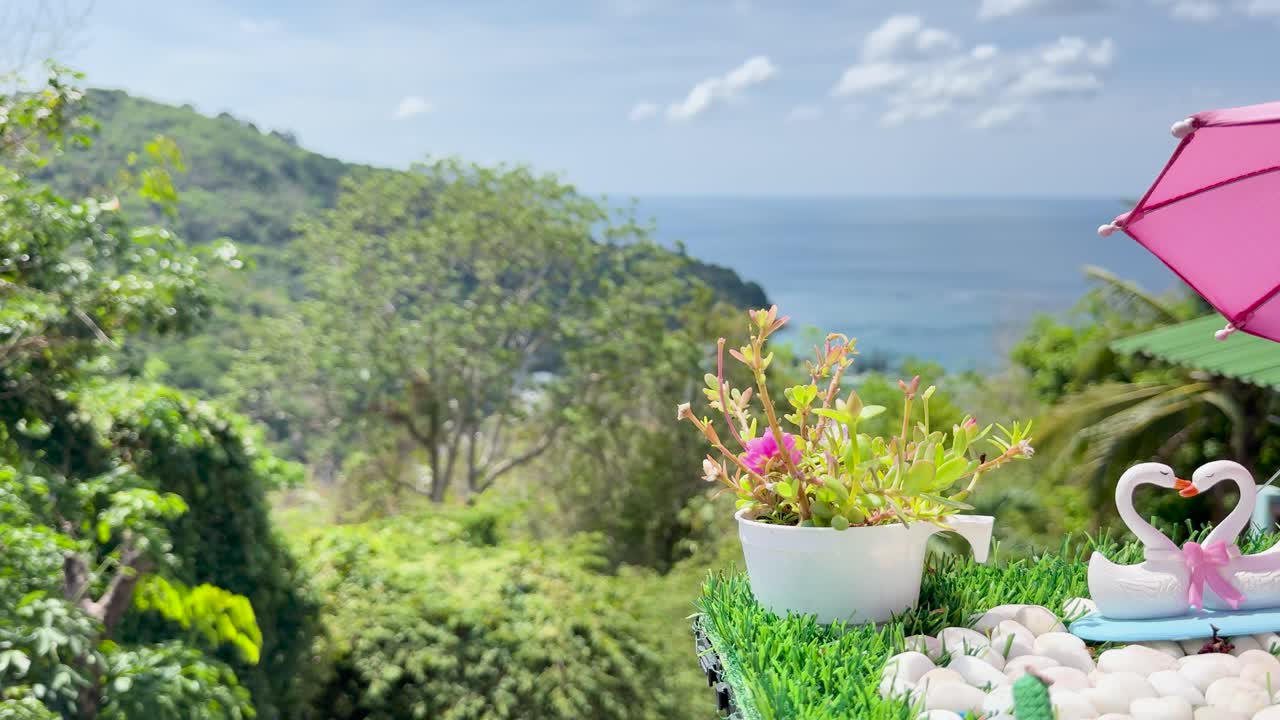 A tranquil clifftop scene in Phuket with decorative swans, vibrant plants, and ocean views under bright daylight