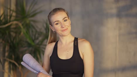 mujer atleta rubia posando con un tapete en la mano y sonriendo mientras está de pie en la habitación del desván