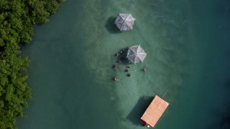 Toma-Aérea-Giratoria-Ascendente-De-Los-Lugareños-Disfrutando-De-Una-Pequeña-Piscina-Turquesa-Natural-Aislada-Con-Una-Ramificación-De-Paraguas-De-Paja-Desde-El-Río-Curimataú-Cerca-De-La-Barra-De-Cunhaú-En-Río-Grande-Do-Norte,-Brasil