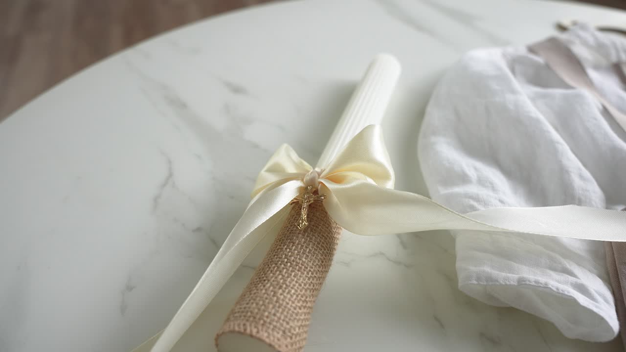 Close-up of a christening candle with a satin bow and burlap handle, laid on a marble surface next to a white linen baby garment, perfect for baptism or ceremonial themes