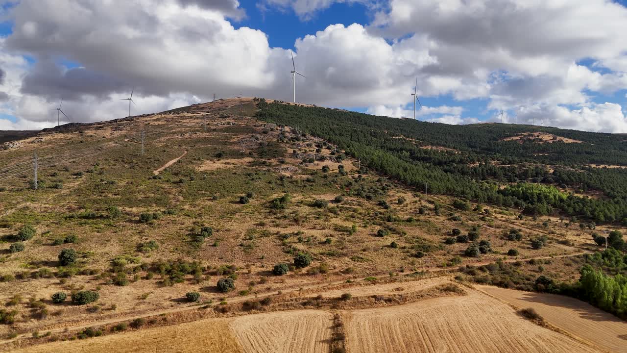Uma tomada aérea suave com movimento dolly avança lentamente em altitude média sobre campos dourados e secos em direção a uma cordilheira de turbinas eólicas, gerando energia verde na paisagem rural de Segóvia.