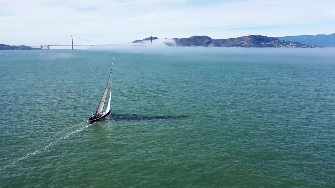 Yksinäinen purjevene purjehtimassa San Franciscon lahdella kohti Golden Gate -siltaa, sumua etäällä. Purjehtii näytön vasemmasta alakulmasta keskelle näyttöä. Paikallaan pysyvä dronekuva.