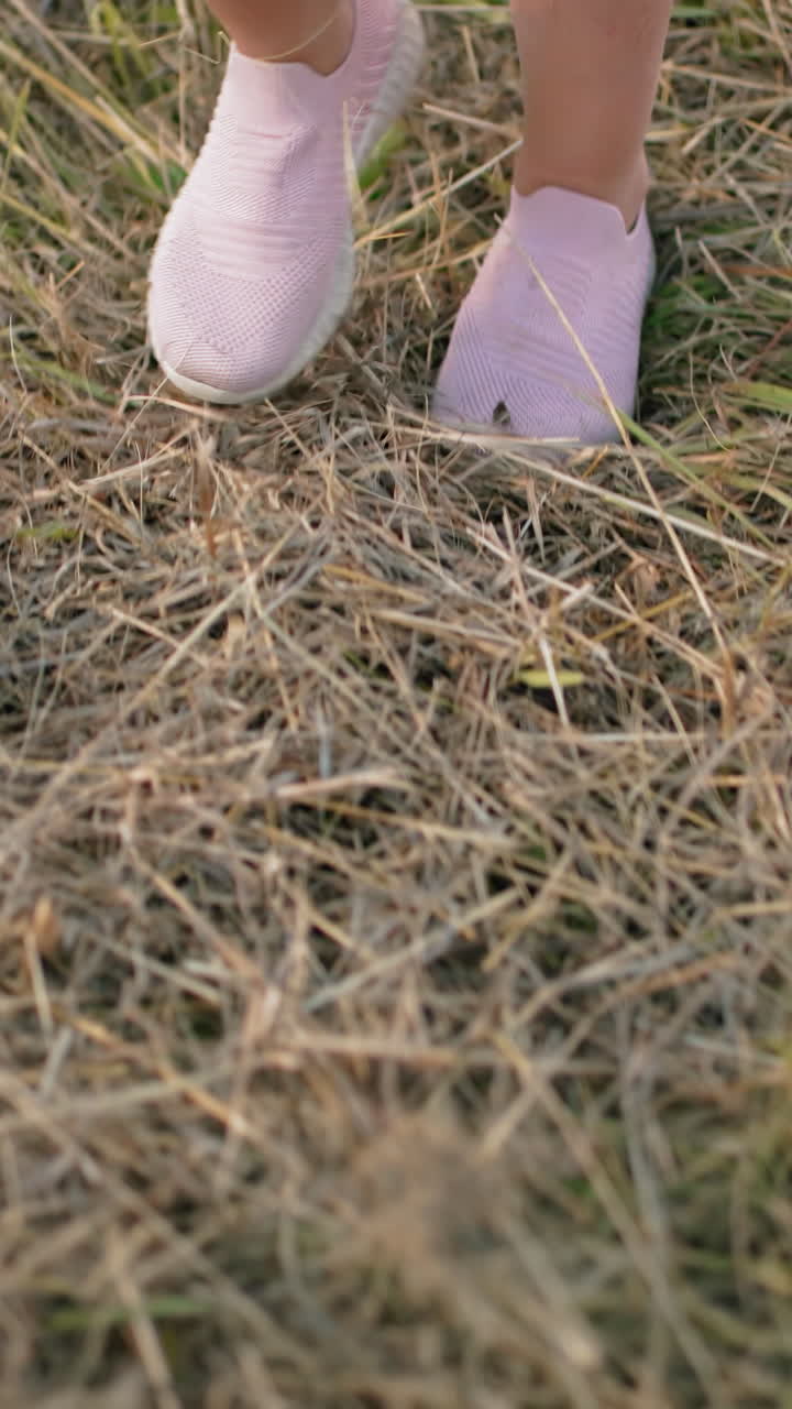 Close-up of person walking on dry grassy field wearing pink slip-on sneakers, steps move naturally over uneven terrain with a mix of green and yellow grass