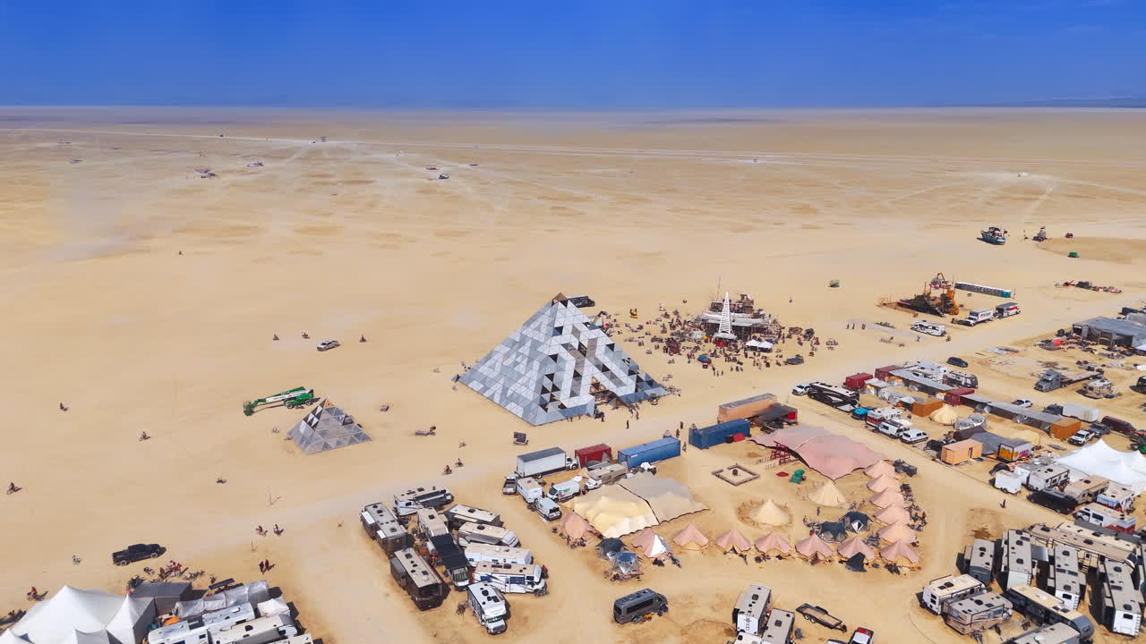 Nevada, USA, 15. August 2025: Fernansicht einer Pyramideninstallation in einer Wüstencamp-Landschaft. Weite Luftaufnahme einer geometrischen Pyramidenstruktur und verschiedener Zelte in einer riesigen, trockenen Wüstenebene.