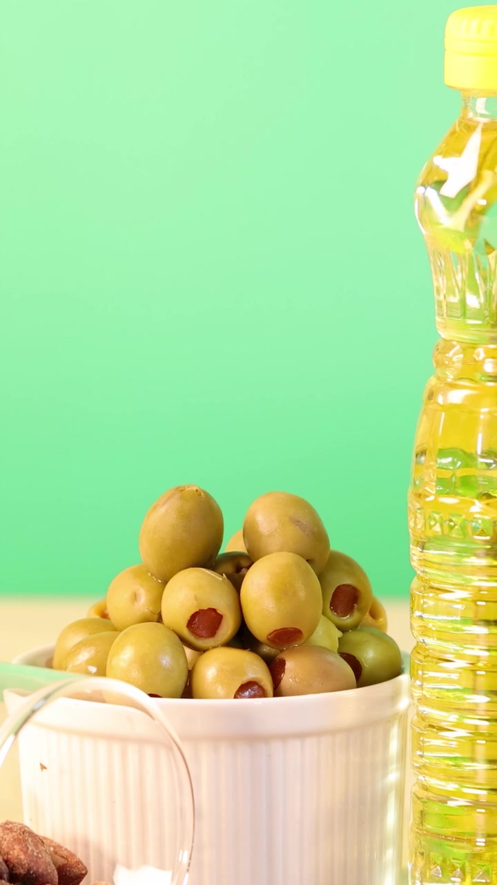Una colorida disposición de aceitunas, almendras y botellas de aceite sobre un fondo verde brillante. Composición estática con iluminación vívida.