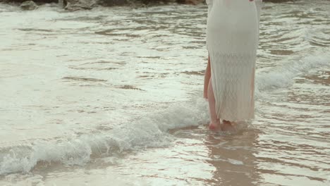 lower half of woman walking in shallow water on beach with gentle waves and white dress