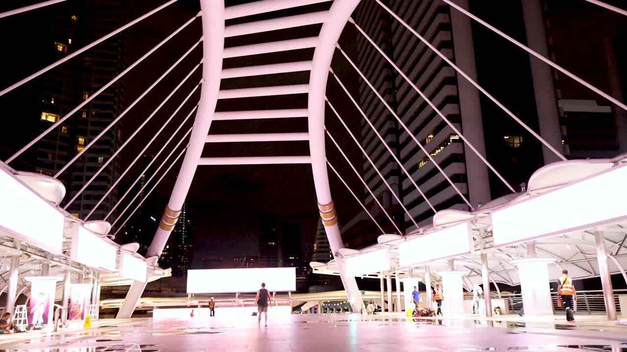 Camera tilts down illuminated Chong Nonsi Bridge, revealing modern architecture and empty urban plaza