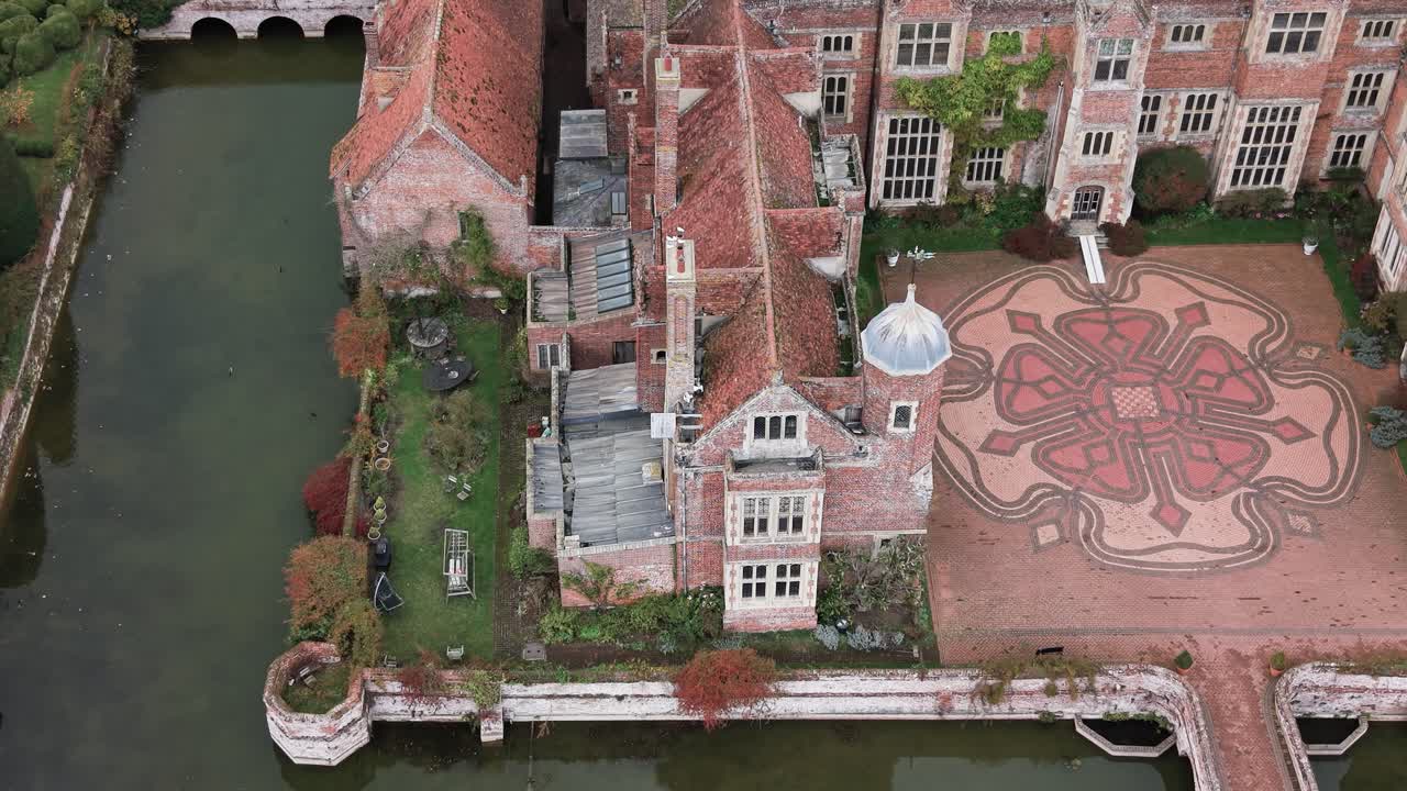 Kentwell Hall, Long Melford, Suffolk United Kingdom - An Old Brick Manor Sits Beside a Calm Moat, Featuring a Detailed Patterned Courtyard and Surrounding Garden - Aerial Drone Shot