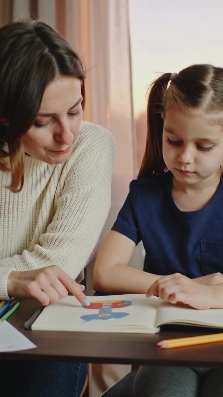 Video verticale: Mamma in maglione chinata che indica, mentre insegna alla figlia in maglietta blu scuro a ricalcare un disegno a casa.
