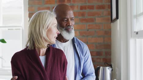 Happy-senior-diverse-couple-in-kitchen,-looking-through-window-and-embracing