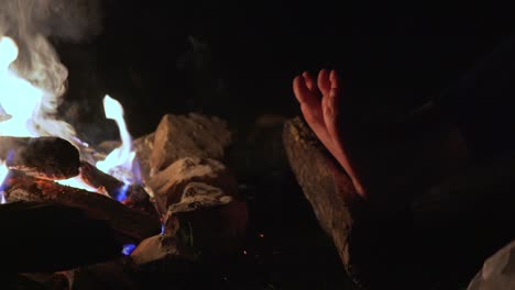 female bask her feet and dance her fingers near a campfire in a night