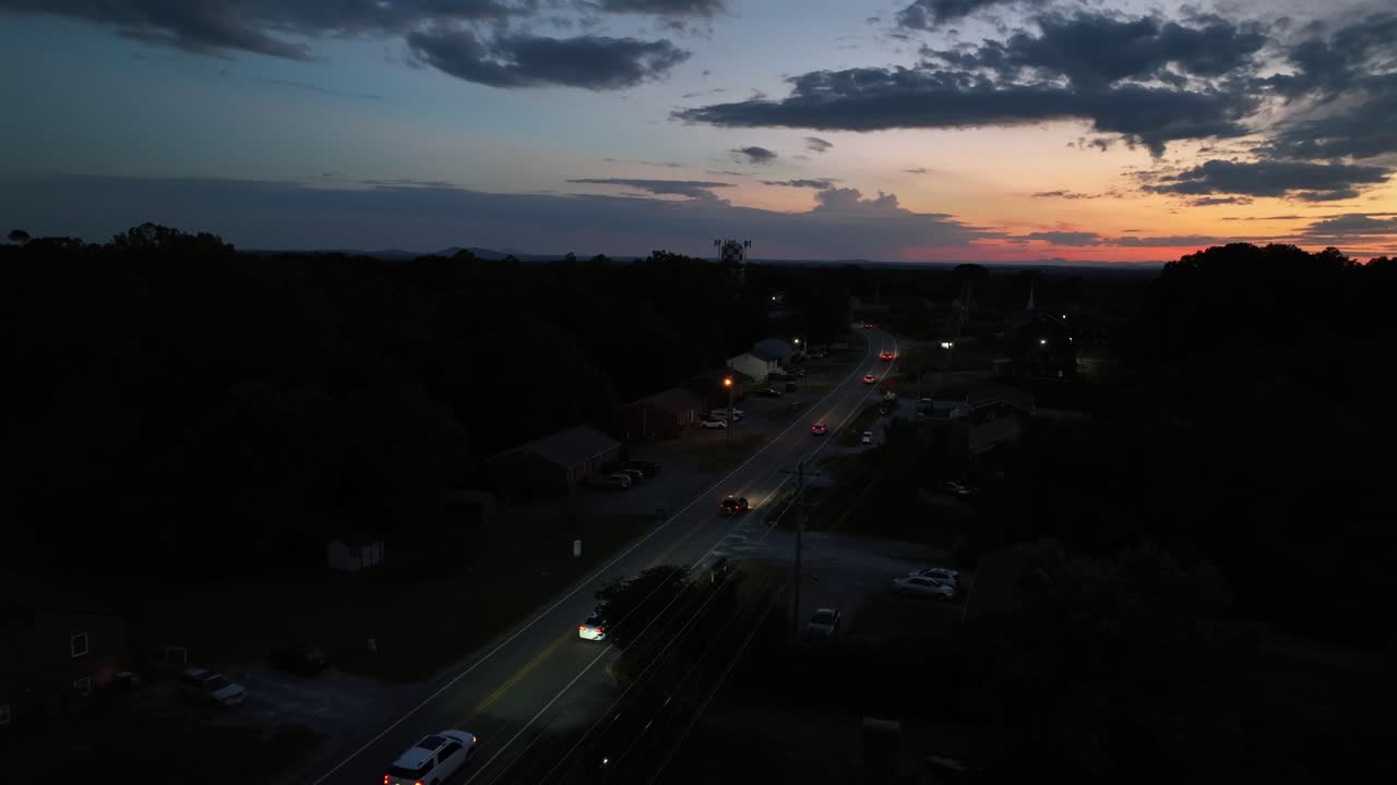 Driving cars on intersection road after golden sunset. Night scene in suburb neighborhood of American. Colored sky at horizon in summer. Aerial wide shot. Headlights of vehicles on street