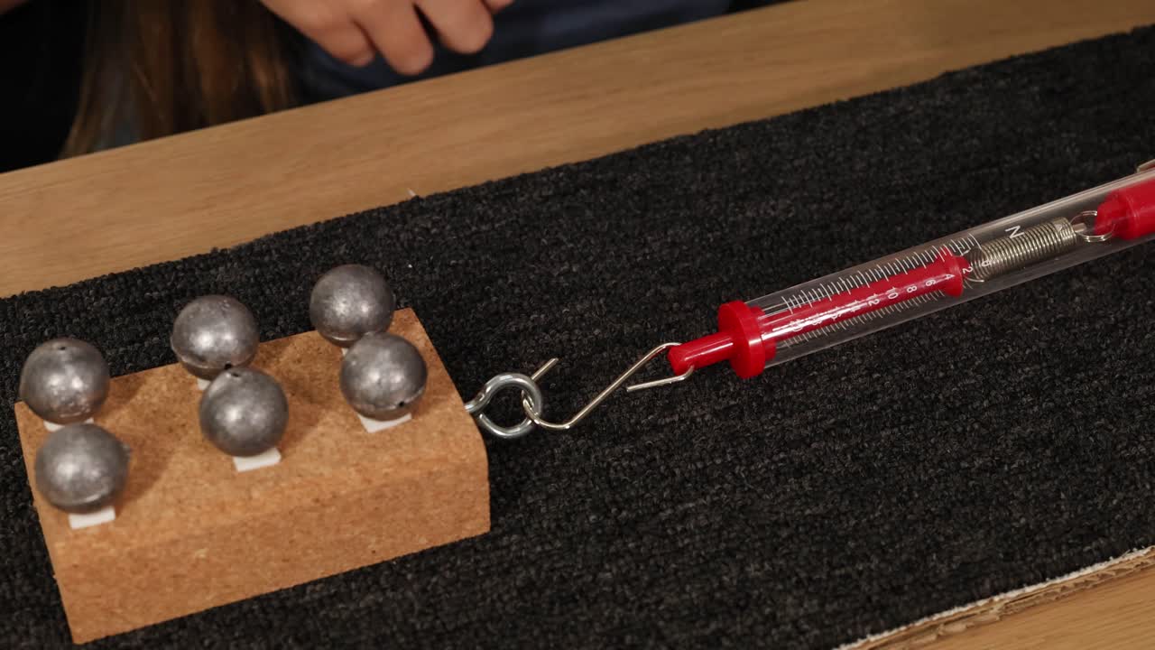 A hand uses a spring scale to measure friction on a carpet, pulling a weighted block across the surface