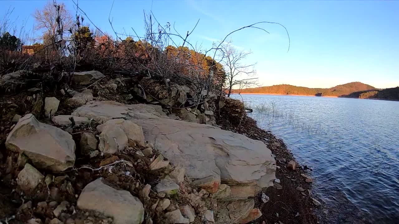 Slow motion view of shoreline of lake with small cliff with rocks and brush with trees in the background and beautiful blue sky
