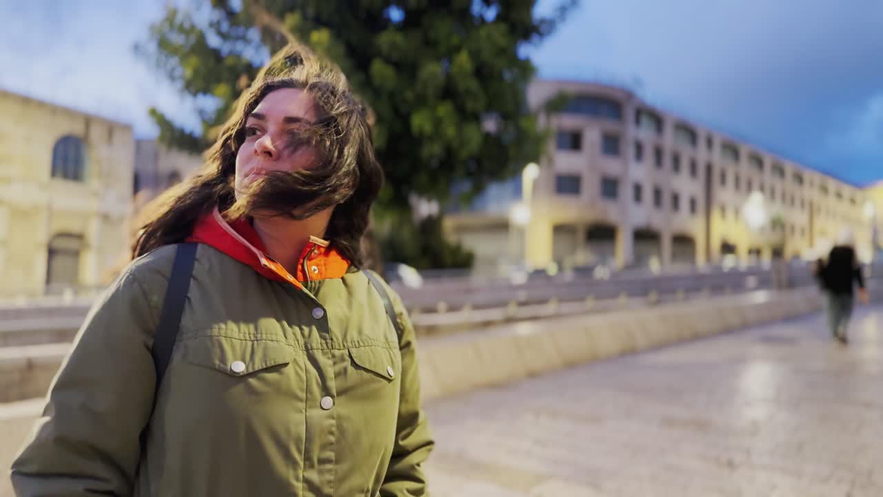 A woman wearing a green jacket and orange scarf stands on a sidewalk