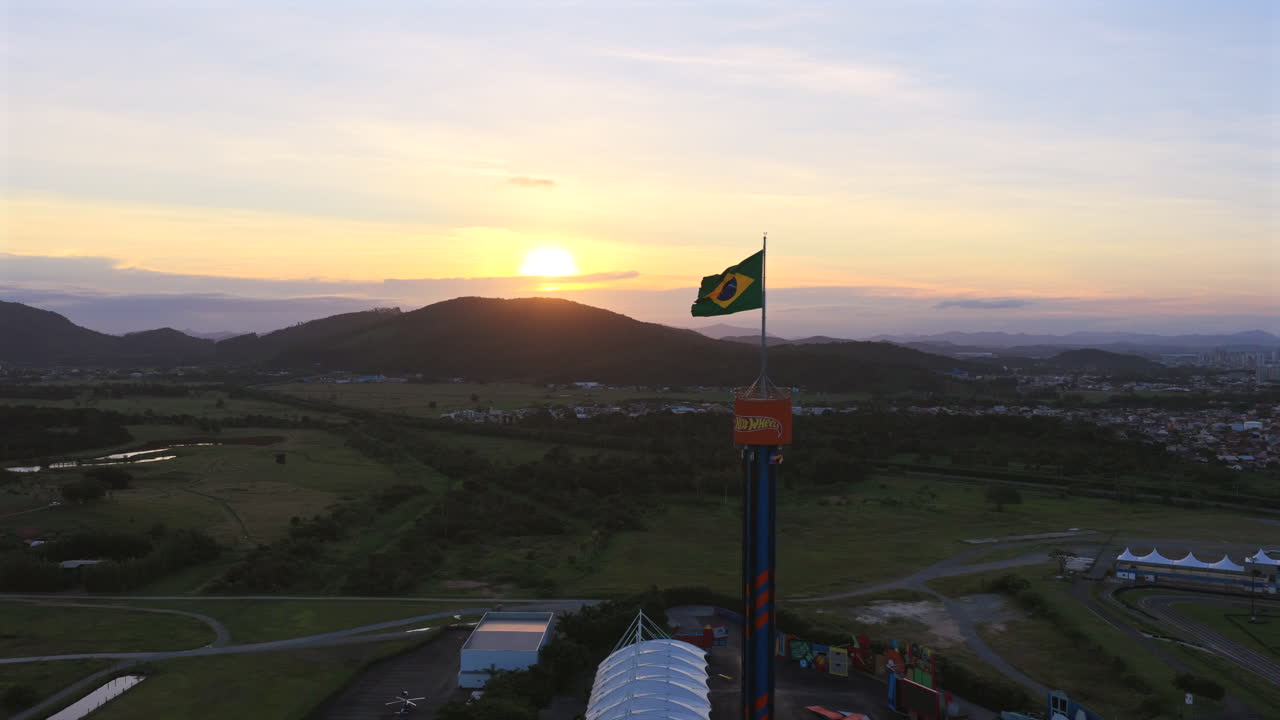 Vista aérea do pôr do sol da bandeira brasileira sobre torre de parque de diversões com paisagem rural no sul do Brasil