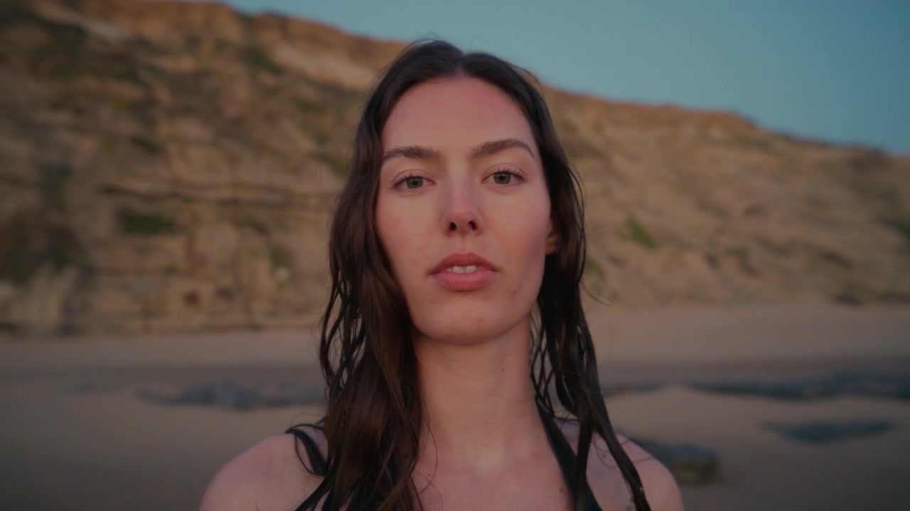 Ritratto ravvicinato di una donna con i capelli bagnati che guarda la fotocamera su una spiaggia sabbiosa vicino a scogliere costiere al tramonto a Ericeira, Portogallo.
