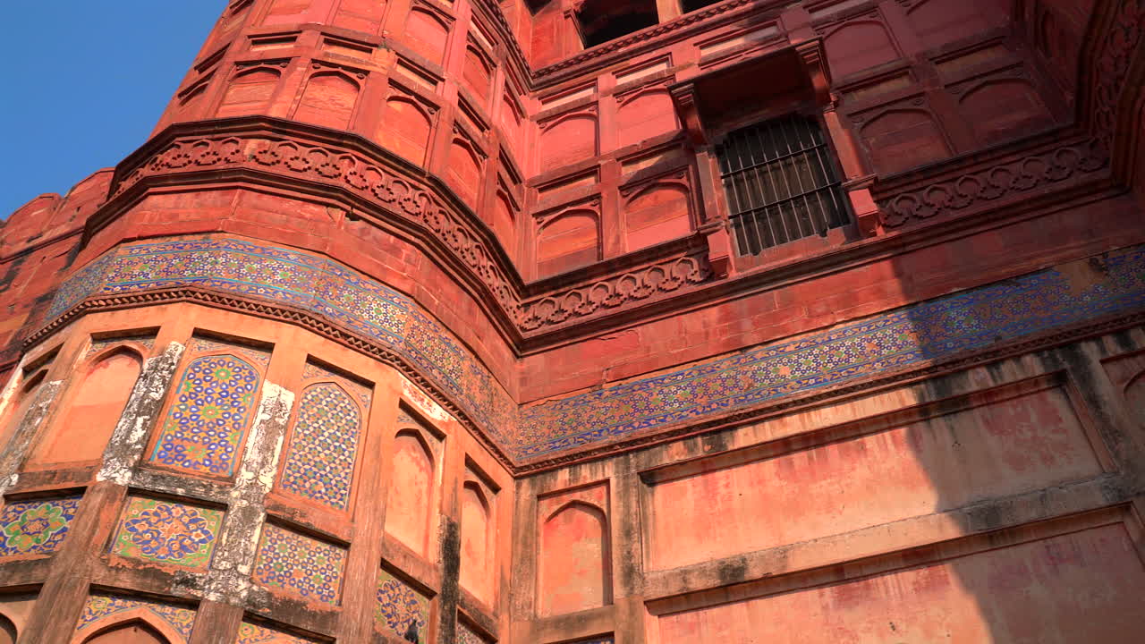 Un'inquadratura statica dal basso degli intricati mosaici di piastrelle blu e gialle e dei dettagli intagliati sulle pareti di arenaria rossa dello storico Forte di Agra nell'Uttar Pradesh, India.