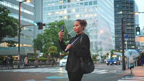 woman stands with a clear umbrella in the evening, using her smartphone, with a backdrop of city lights