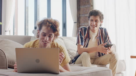 Happy-diverse-gay-male-couple-sitting-at-sofa-using-tablet-and-smartphone-at-home,-slow-motion