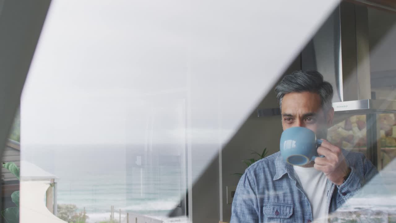 feliz hombre biracial bebiendo taza de café y pensando en la cocina solo