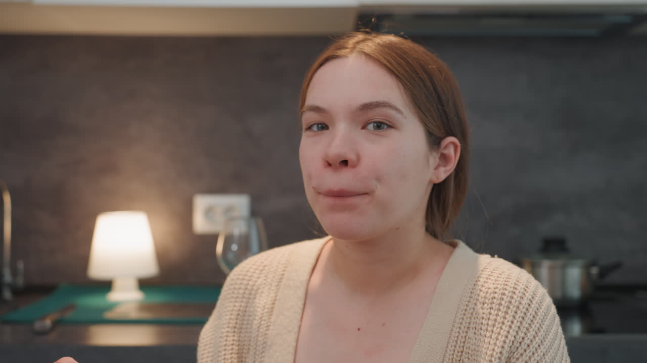 Pregnant white woman smiling after tasting in kitchen, sampling homemade breakfast, glass and stove visible, satisfied playful expression, beige cardigan, lamp glow, casual morning routine, prenatal