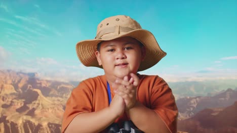 asian boy with a hat clapping hands after looking through the binoculars. boy researcher examines something while traveling at the top of mountain, travel tourism adventure concept, close up