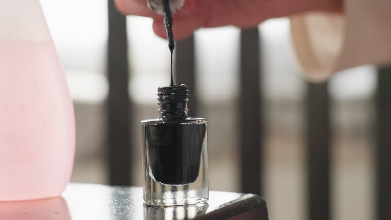 Close up student hands lift white cap from black nail polish bottle while drop drips back into glass, brush held over rim, manicure prep on table with pink remover nearby, focus on hygiene