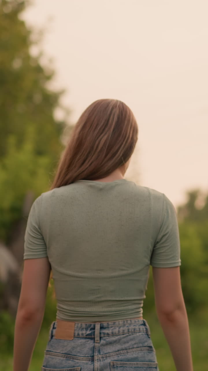 잔디로 된 길을 따라 걸어가는 젊은 여성, 그녀의 머리카락이 앞으로 흐르고, 전선 에서 조용하고 자연스러운 장면을 만들고, 먼 차와 초록색을 특징으로하는 흐릿한 배경