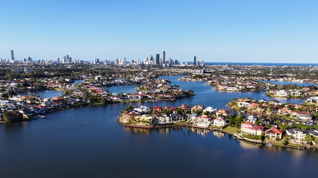 Drone camera glides smoothly above Gold Coast riverfront neighborhoods, revealing luxury homes, calm water, and city skyline under bright daylight