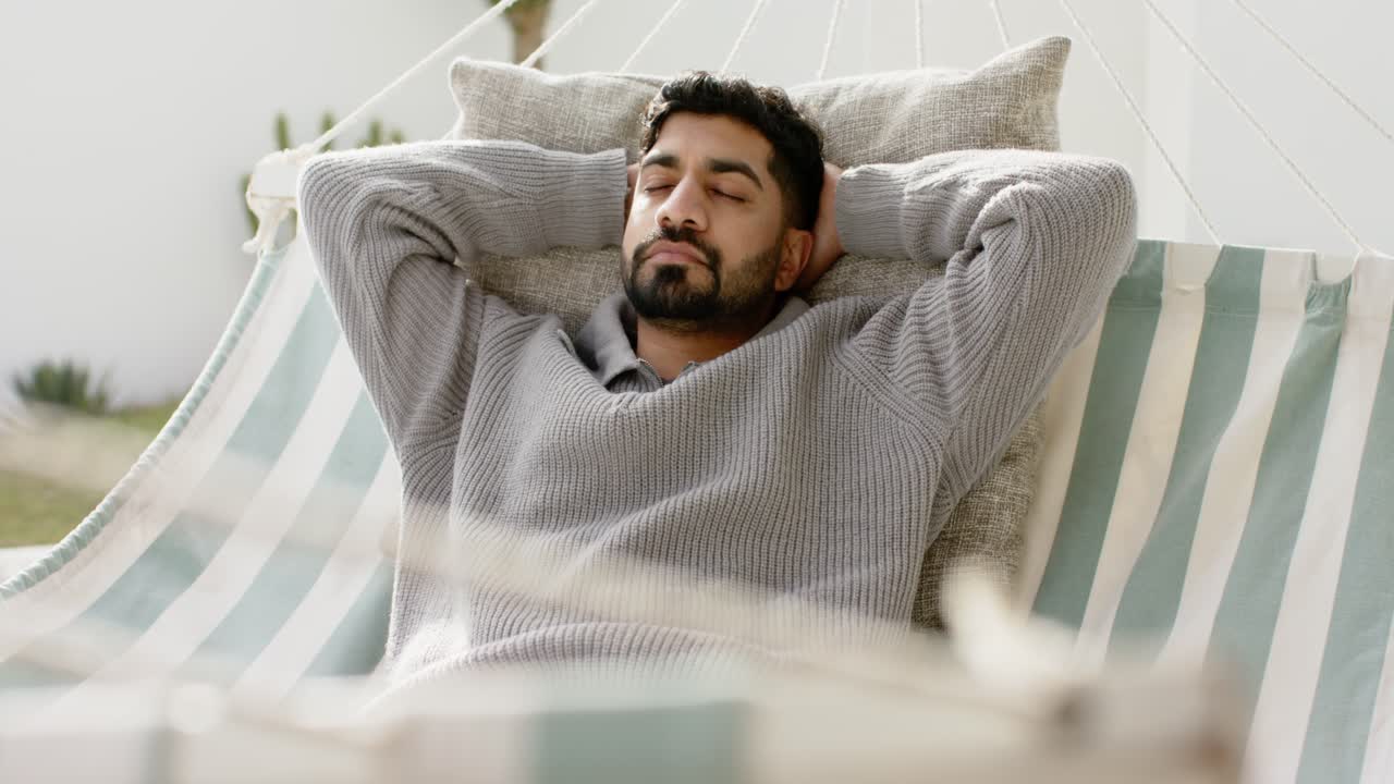 Uomo indiano adulto disteso su un cuscino, che chiude gli occhi e respira in un'amaca a righe su un patio illuminato dal sole