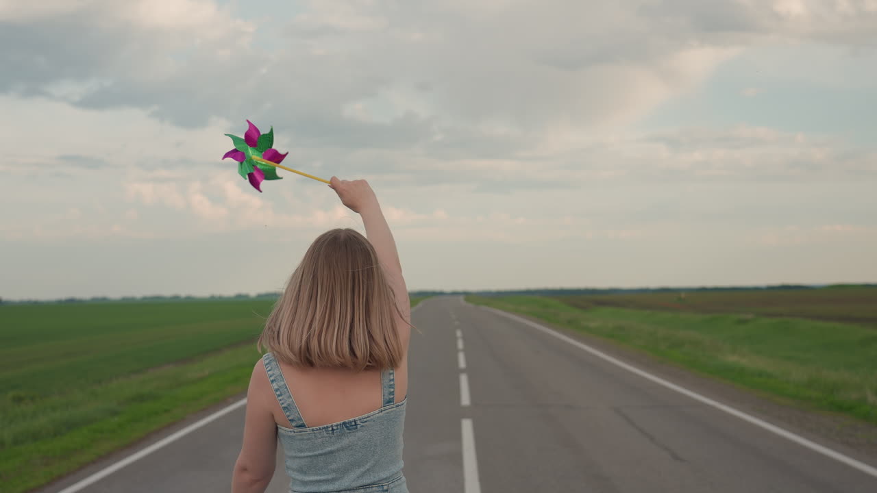 Back view of woman waving colourful pinwheel while walking along straight asphalt road bordered by green fields under cloudy sky hair swaying in breeze portraying whimsical journey motion joy